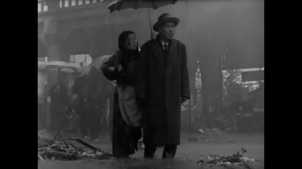 A still from the movie Ikiru. The main character, a middle aged man stands in water with his hat and coat on. A woman, dressed poorly, stands behind him, holding an umbrella over his head.