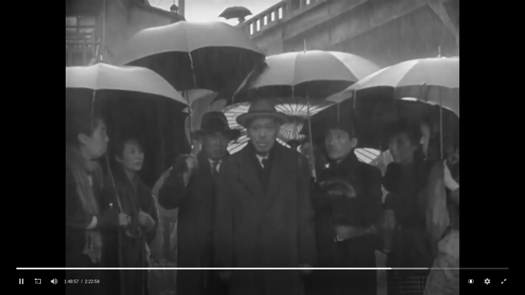 A still from the movie Ikiru. The main character, a middle aged man stands with his hat and coat on. Two of his colleagues, similarly dressed, stand beside him with umbrellas. Local woman stand on either side, also holding umbrellas.